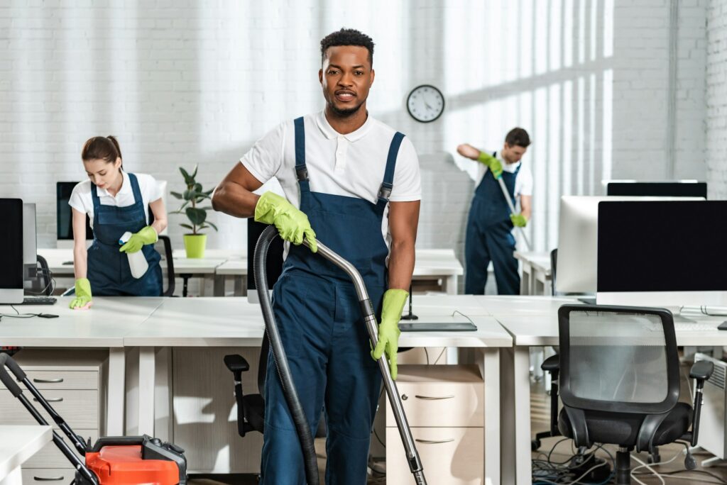 handsome african american cleaner looking at camera while standing with vacuum cleaner near